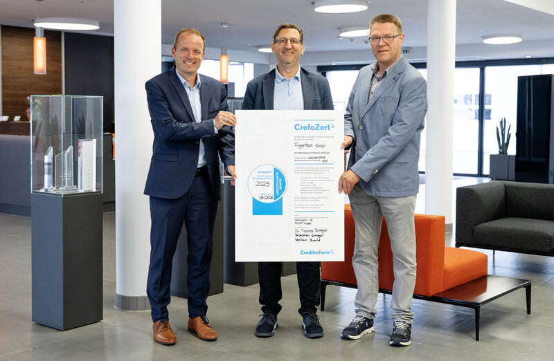 Drei Männer in Businesskleidung stehen in einem modernen Büro und halten gemeinsam eine große Urkunde mit dem Titel 'CrefoZert'