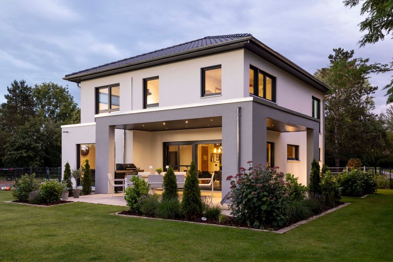 Zweistöckiges, modernes Wohnhaus mit weißer Fassade, Flachdach mit leichter Neigung, überdachter Terrasse und großen Fensterflächen, umgeben von gepflegtem Garten und Rasenfläche bei Dämmerung.