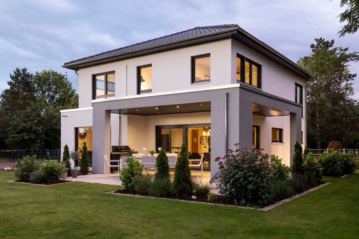 Zweistöckiges, modernes Wohnhaus mit weißer Fassade, Flachdach mit leichter Neigung, überdachter Terrasse und großen Fensterflächen, umgeben von gepflegtem Garten und Rasenfläche bei Dämmerung.