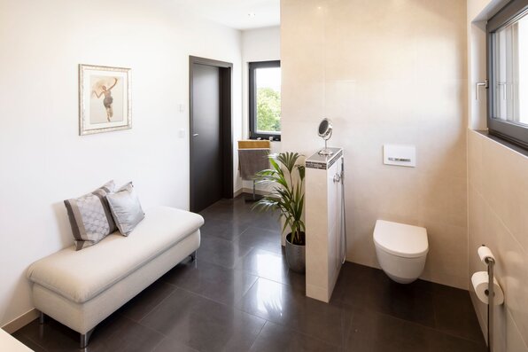 Modernes Badezimmer in einem frei geplanten FingerHaus mit Wand-WC, großformatigen Fliesen und heller Wandgestaltung. Sitzbank mit Kissen und dekorative Pflanzen schaffen wohnliches Ambiente.