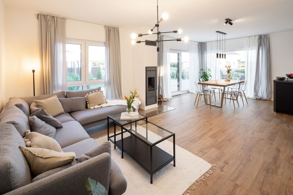 Offener Wohn- und Essbereich mit grauer Eckcouch, schwarzem Couchtisch und Esstisch im Hintergrund. Große Fenster, helle Wände und Holzboden schaffen eine warme Atmosphäre.
