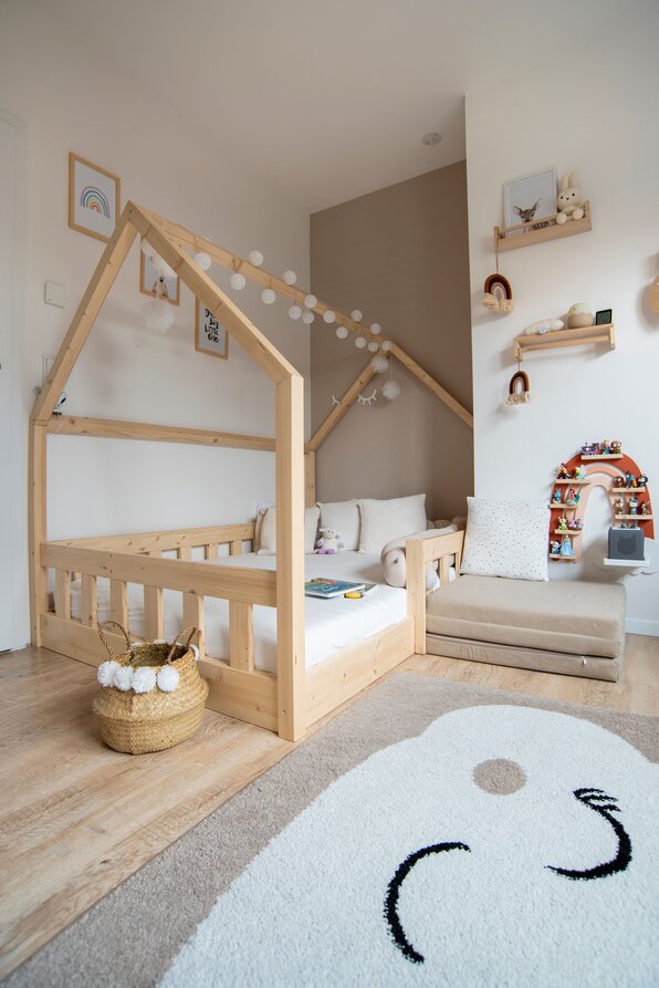 Kinderzimmer mit Hausbett aus Holz, hellem Teppich, rundem Sitzkissen und dezenten Wandregalen.