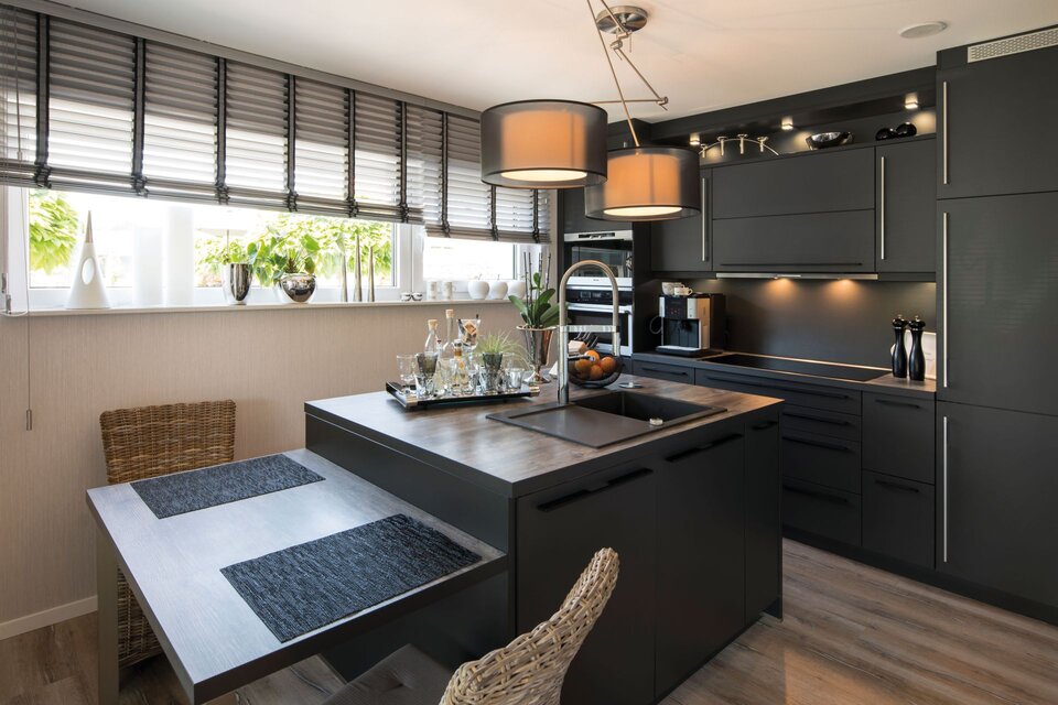 Moderne Küche mit dunklen Fronten, integrierter Sitzecke und Holzarbeitsplatte. Große Fenster mit Jalousien, zwei Deckenlampen und Pflanzen verleihen dem Raum eine warme Atmosphäre.