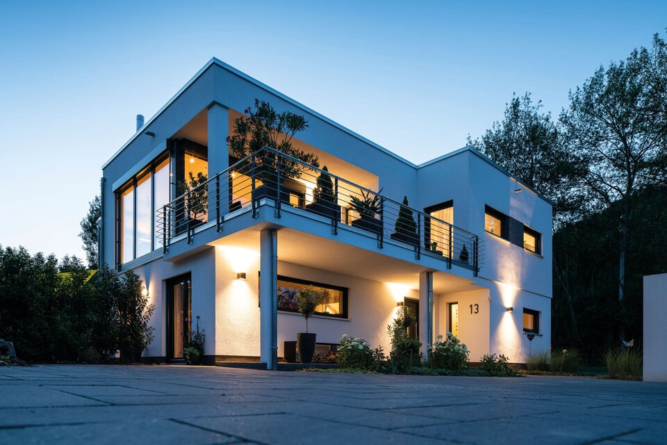 Modernes FingerHaus im Bauhausstil bei Abenddämmerung mit Flachdach, beleuchtetem Eingangsbereich, großem Balkon und bodentiefen Fenstern.