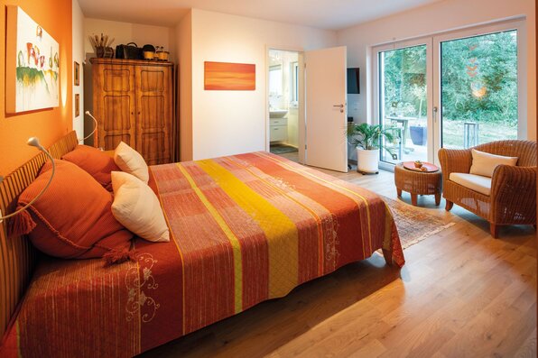 Gemütliches Gästezimmer in einem frei geplanten FingerHaus mit Doppelbett in warmen Farben, Holzschrank, Sitzgruppe und Zugang zur Terrasse durch große Fenster.