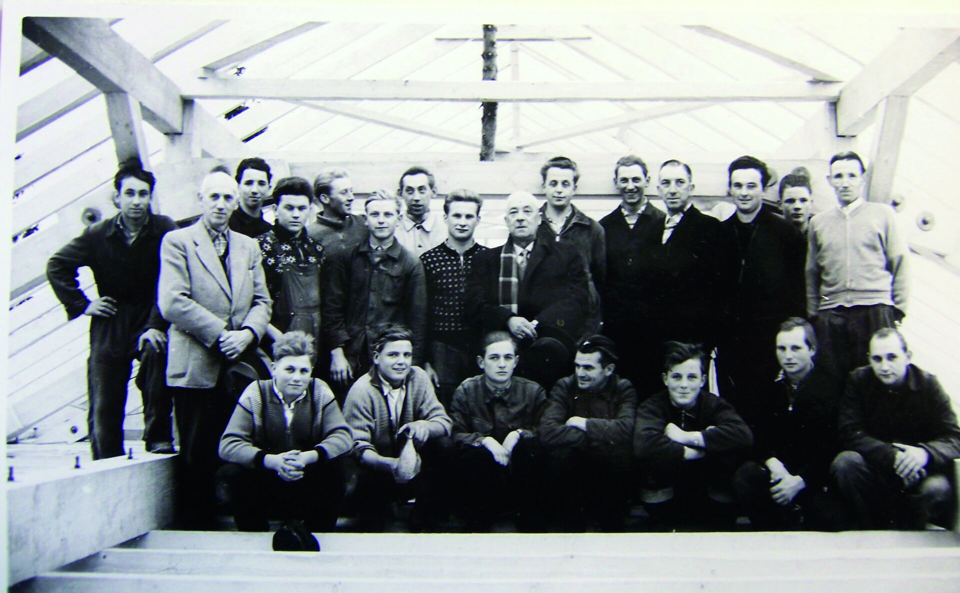 Gruppe von Personen in einem Holzrahmenbau mit sichtbaren Balken und Dachkonstruktion, einige sitzen, andere stehen, alle in lässiger Kleidung.