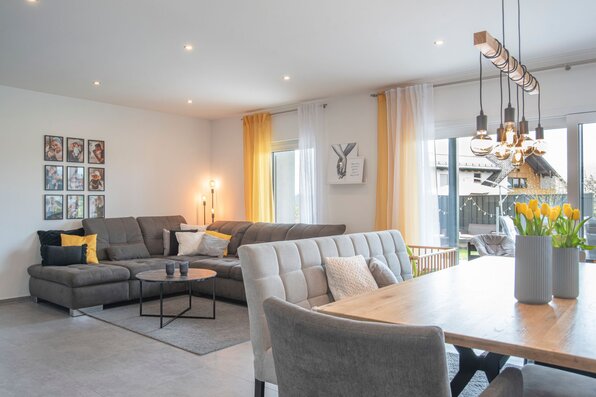 Heller Wohn- und Essbereich in einem FingerHaus NIVO 130 mit großer grauer Eckcouch, Esstisch aus Holz, modernen Lampen, gelben Dekoelementen und Blick in den Garten.