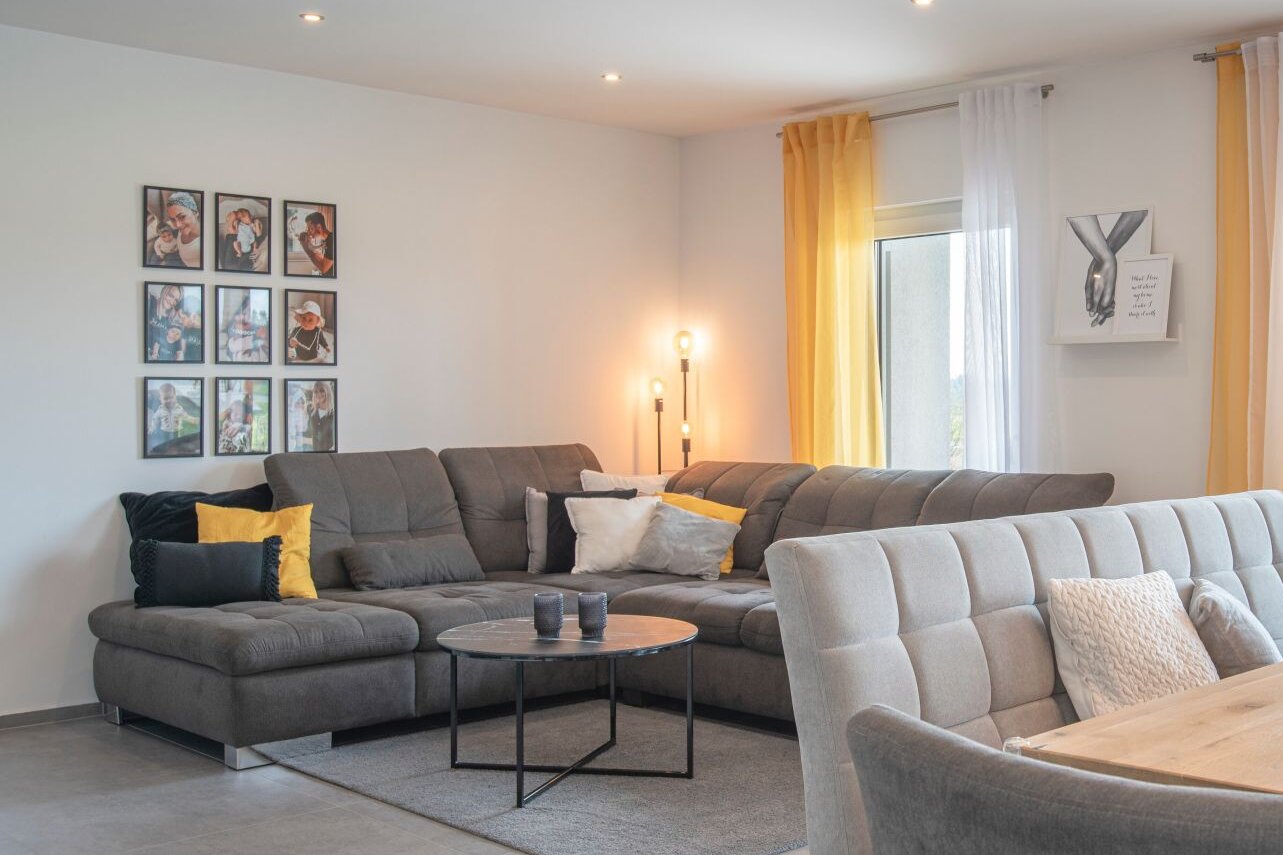 Heller Wohn- und Essbereich in einem FingerHaus NIVO 130 mit großer grauer Eckcouch, Esstisch aus Holz, modernen Lampen, gelben Dekoelementen und Blick in den Garten.