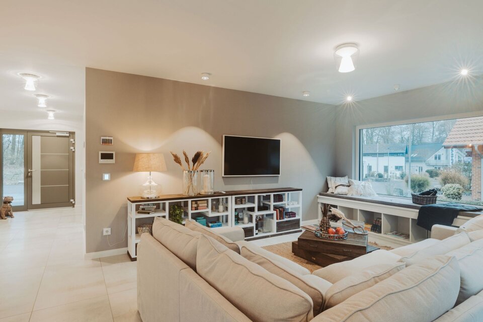 Wohnzimmer mit heller Eckcouch, großem Fenster und TV-Wand mit offener Regalablage im Musterhaus SENTO Bad Vilbel. Holz-Couchtisch, helle Fliesen und dezente Beleuchtung schaffen eine warme, moderne Atmosphäre.