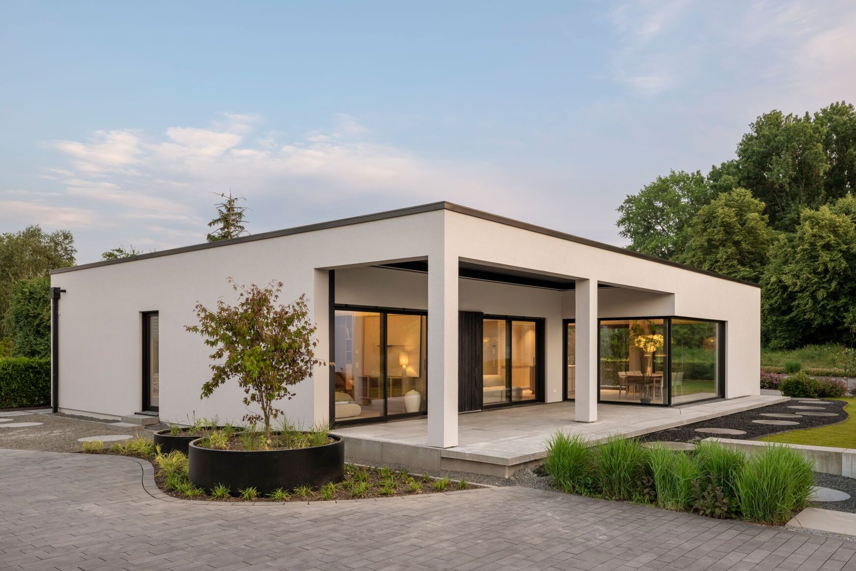 Musterhaus NIVO in Frankenberg mit moderner Bungalowarchitektur, weißer Putzfassade, überdachter Terrasse und großzügigen Eckfenstern in der Abenddämmerung.