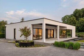 Musterhaus NIVO in Frankenberg mit moderner Bungalowarchitektur, weißer Putzfassade, überdachter Terrasse und großzügigen Eckfenstern in der Abenddämmerung.