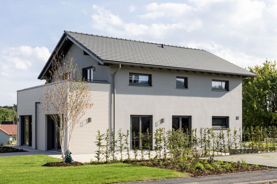 Moderne Außenansicht des FingerHaus ARTIS 300 Musterhauses Würzburg mit grauer Putzfassade, Satteldach und vorgelagerter Holzverkleidung.