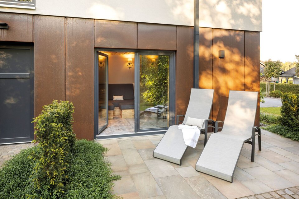 Terrasse eines modernen FingerHaus-Hauses mit zwei stilvollen Liegestühlen vor bodentiefen Fenstern, die in einen gemütlich beleuchteten Saunabereich führen.