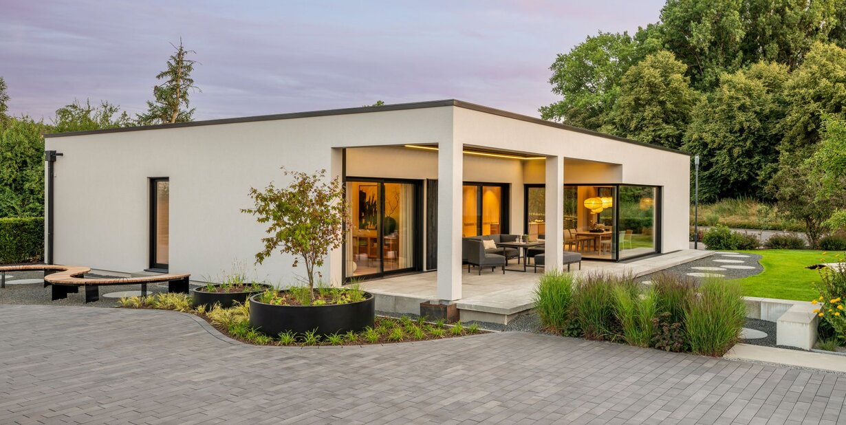 FingerHaus Musterhaus NIVO in Frankenberg, moderner Bungalow mit Flachdach, überdachter Terrasse und großen Fensterflächen in der Abenddämmerung.