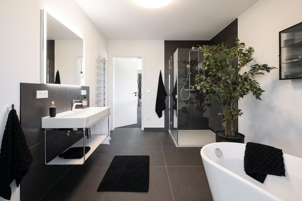 Großzügiges Badezimmer im Musterhaus TALO Fulda mit freistehender Badewanne, moderner Dusche, Waschtisch und eleganten schwarzen Akzenten.