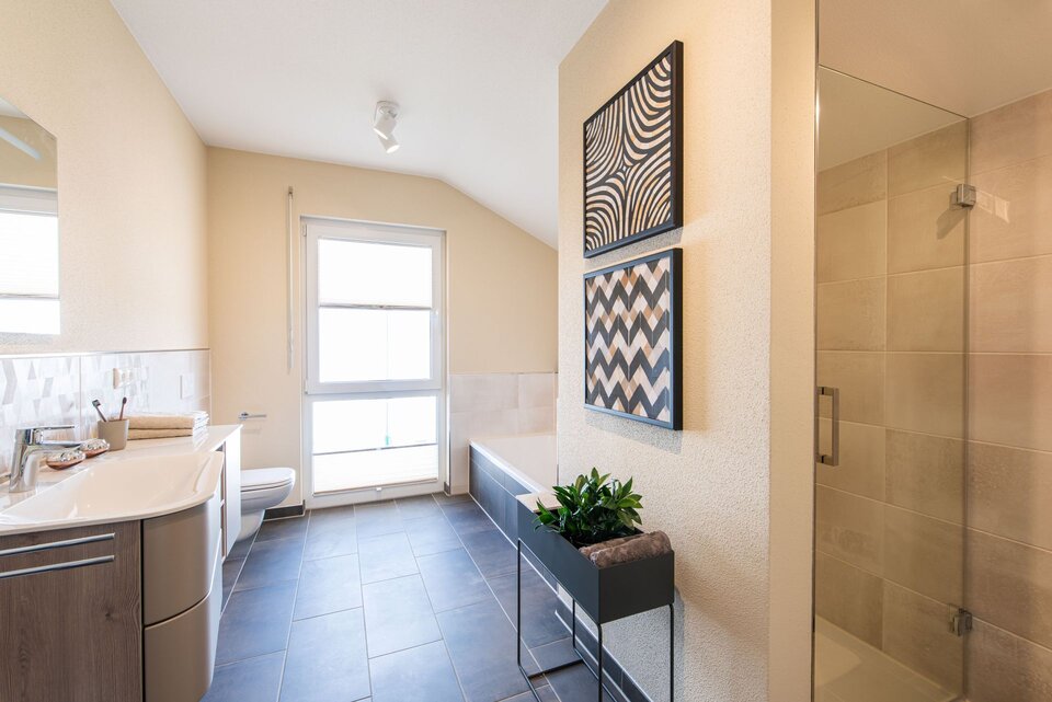Modernes Badezimmer im FingerHaus Musterhaus TALO in Babenhausen mit Dusche, Badewanne, Waschtisch und dekorativen Wandbildern. Helle Fliesen kombiniert mit dunklem Bodenbelag und stilvollen Einrichtungselementen.