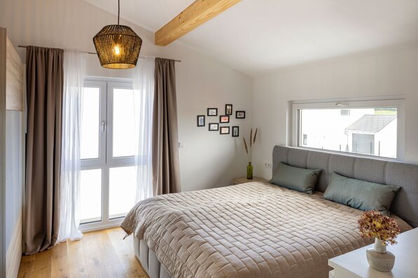 Schlafzimmer mit großem Doppelbett, gepolstertem Kopfteil, bodentiefem Fenster, breitem Querfenster und sichtbarem Holzbalken unter der Dachschräge.