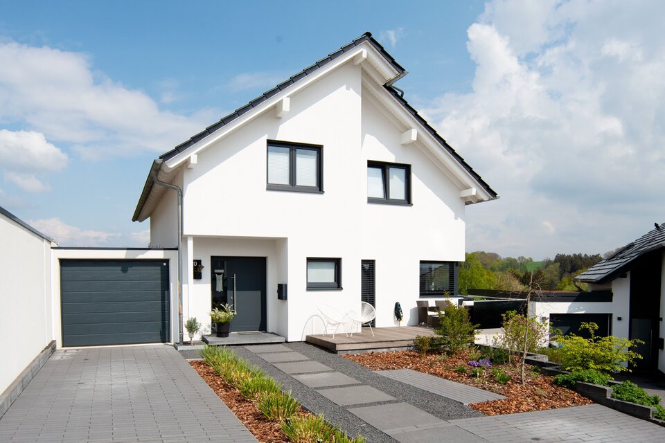 Modernes frei geplantes FingerHaus mit weißer Fassade, dunklem Satteldach und Garage, Blick auf die Einfahrt und Vorgarten.