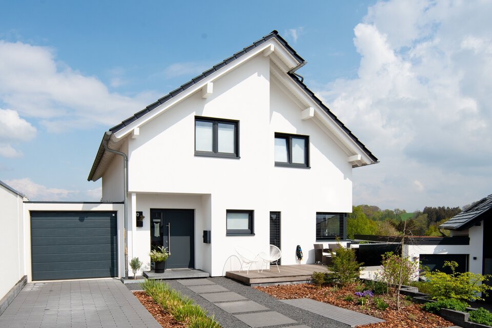 Modernes frei geplantes FingerHaus mit weißer Fassade, dunklem Satteldach und Garage, Blick auf die Einfahrt und Vorgarten.