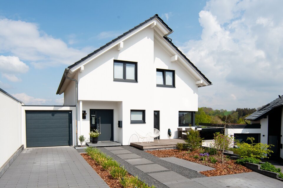 Modernes frei geplantes FingerHaus mit weißer Fassade, dunklem Satteldach und Garage, Blick auf die Einfahrt und Vorgarten.
