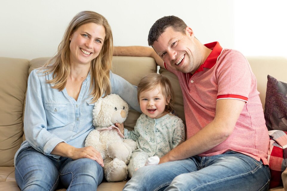 Glückliche Familie sitzt gemeinsam auf dem Sofa im FingerHaus Wohnzimmer, mit Kind und Kuscheltier.