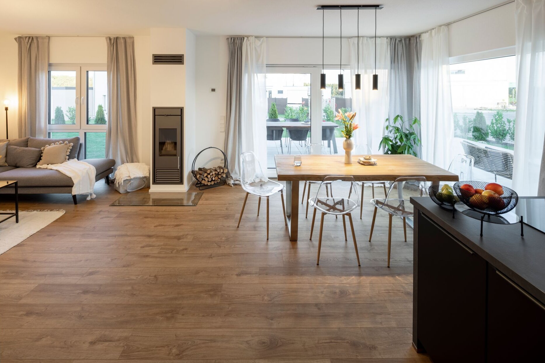 Lichtdurchfluteter Wohn- und Essbereich mit großem Holztisch, Kaminofen und bodentiefer Fensterfront; moderne und gemütliche Atmosphäre.