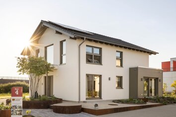 Außenansicht des FingerHaus Musterhauses ARTIS 300 in Nürnberg mit weißer Putzfassade, dunkel gedecktem Satteldach und seitlichem Anbau mit horizontaler Fassadenverkleidung; davor gepflasterter Weg, Hochbeete mit Bepflanzung, Bäume, Fahnenmasten und tief stehende Sonne.