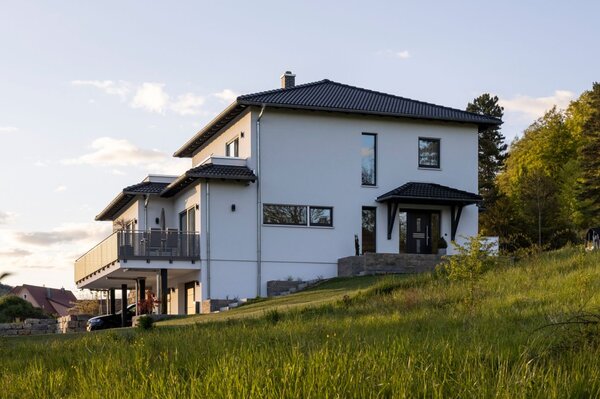 Modernes frei geplantes FingerHaus mit weißer Fassade, großem Balkon und Hanglage im Grünen bei Sonnenuntergang.