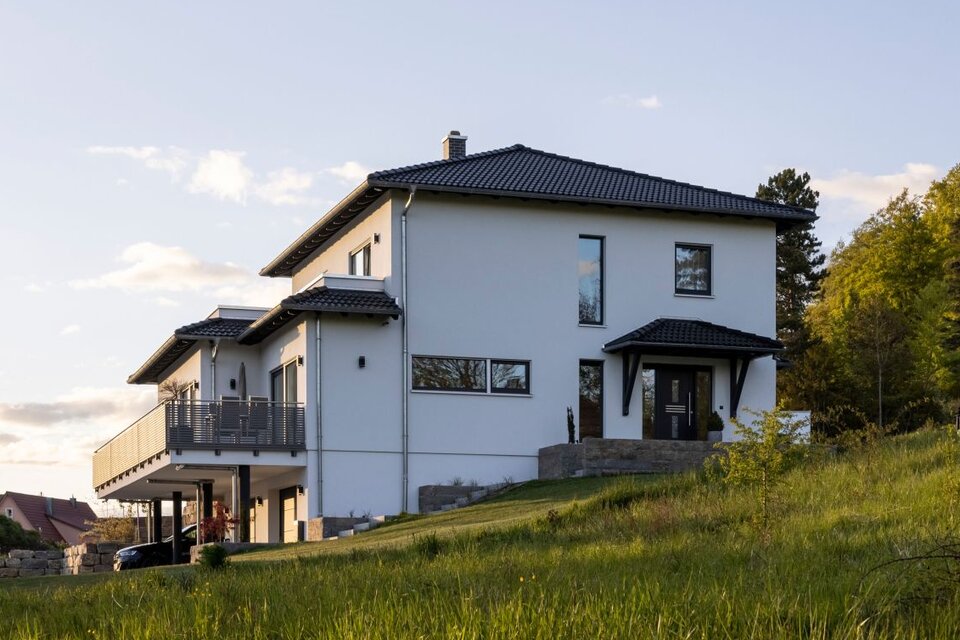 Modernes frei geplantes FingerHaus mit weißer Fassade, großem Balkon und Hanglage im Grünen bei Sonnenuntergang.