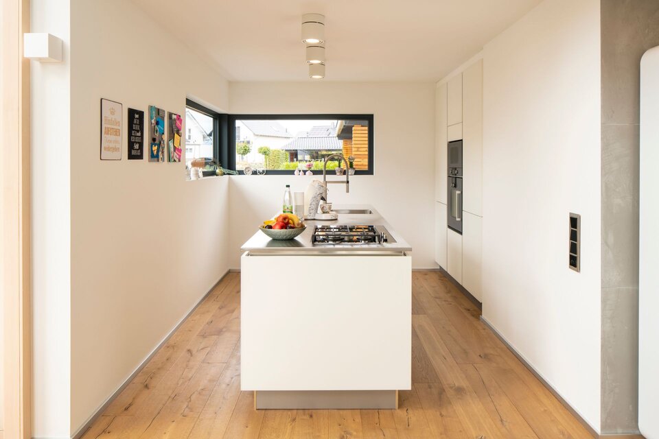 Moderne weiße Küche mit großzügiger Arbeitsinsel, Holzboden und viel Tageslicht.