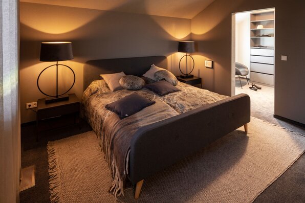 Schlafzimmer im FingerHaus ARTIS 400 Musterhaus Schwarzwald mit grauem Doppelbett, stimmungsvoller Beleuchtung, Teppichboden und direktem Zugang zum Ankleidezimmer.