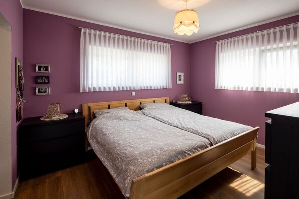 Schlafzimmer mit violetten Wänden, Doppelbett aus Holz, großen Fenstern und weißen Vorhängen
