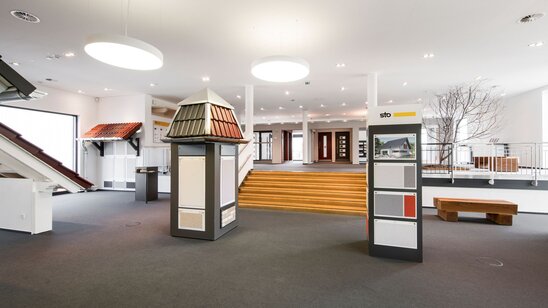 Innenaufnahme der FingerHaus-Bemusterung mit Ausstellungsflächen für Dachziegel, Fassaden, Farben und Materialien. Der Raum ist hell gestaltet, mit großen Leuchten, Treppen und verschiedenen Musterpräsentationen.