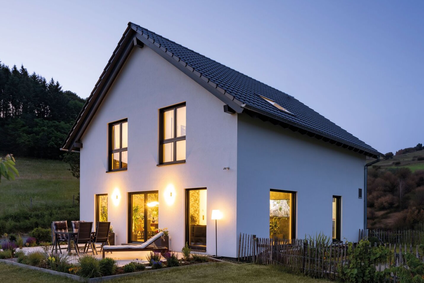 Zweigeschossiges Einfamilienhaus von FingerHaus in der Abenddämmerung mit beleuchteten Fenstern, Garten und Terrasse. Das Haus steht am Waldrand mit Blick auf eine hügelige Landschaft.
