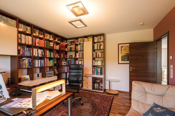 Arbeitszimmer mit großem Bücherregal, modernem Schreibtisch und Lederstuhl auf einem Teppichboden mit Holzakzenten.