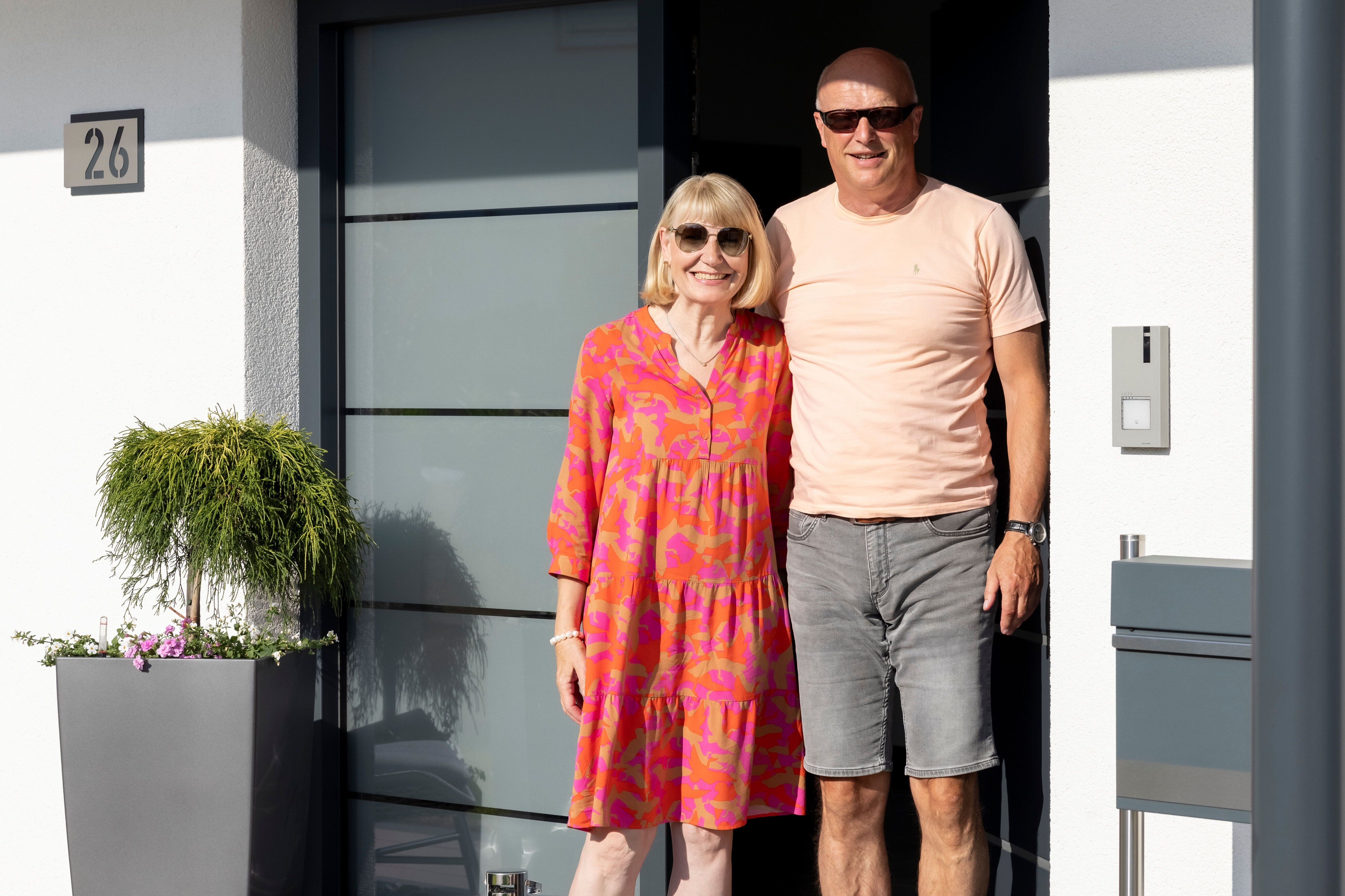Zwei Personen stehen vor der Haustür eines modernen Fertighauses von FingerHaus mit Hausnummer 26, links eine große Pflanzvase und mehrere Laternen auf der Veranda.