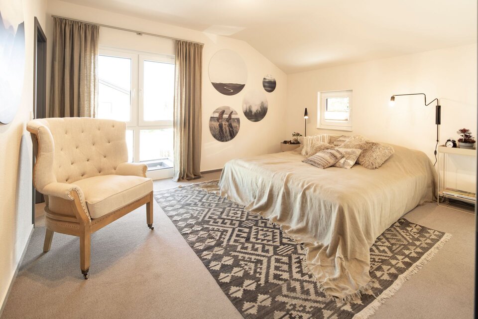 Helles Schlafzimmer mit Doppelbett, beigem Sessel und großem Fenster. Wandbilder in Rundformen, Teppich mit Muster und helle Stoffe schaffen eine warme, ruhige Atmosphäre.