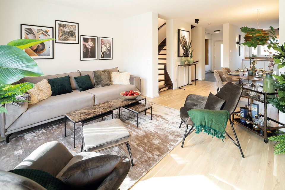 Wohnzimmer mit hellem Sofa, dunklem Sessel, Teppich und Bildern; im Hintergrund Flur und Eingang.