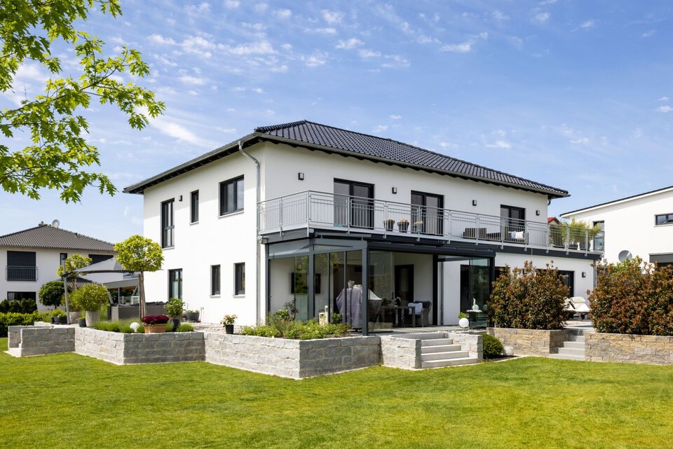 Modernes Zweifamilienhaus von FingerHaus mit Balkon, überdachter Terrasse und weitläufigem Garten an einem sonnigen Tag.
