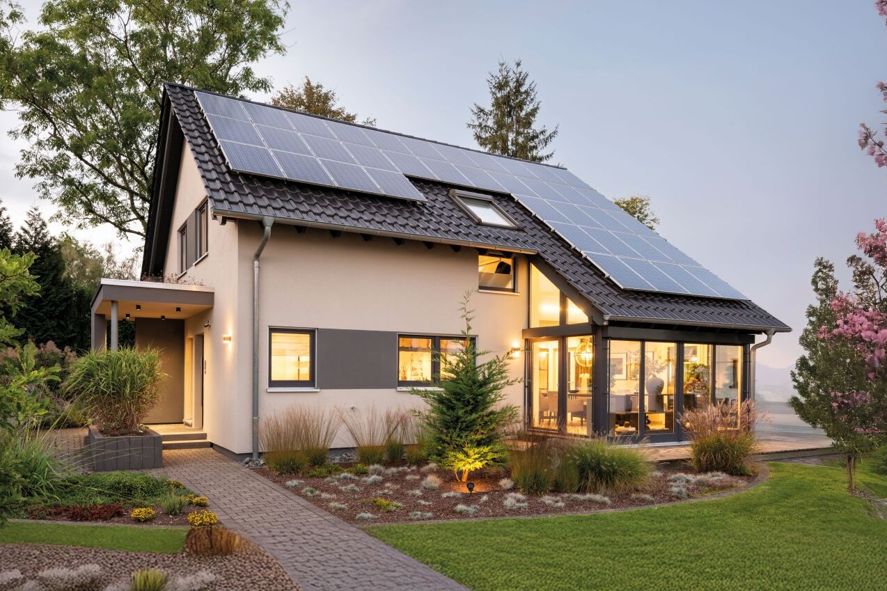 Zweistöckiges Wohnhaus mit hellem Putz, geneigtem Satteldach mit Photovoltaikmodulen, großen Fensterflächen, verglastem Anbau, Hauseingang mit Vordach und angelegtem Garten bei Dämmerung.