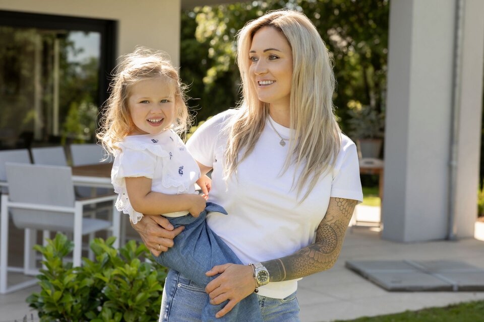 Eine Frau hält ihre lachende Tochter im Arm, beide stehen auf der sonnigen Terrasse eines modernen FingerHaus-Hauses, umgeben von grünen Pflanzen und moderner Gartenmöblierung.