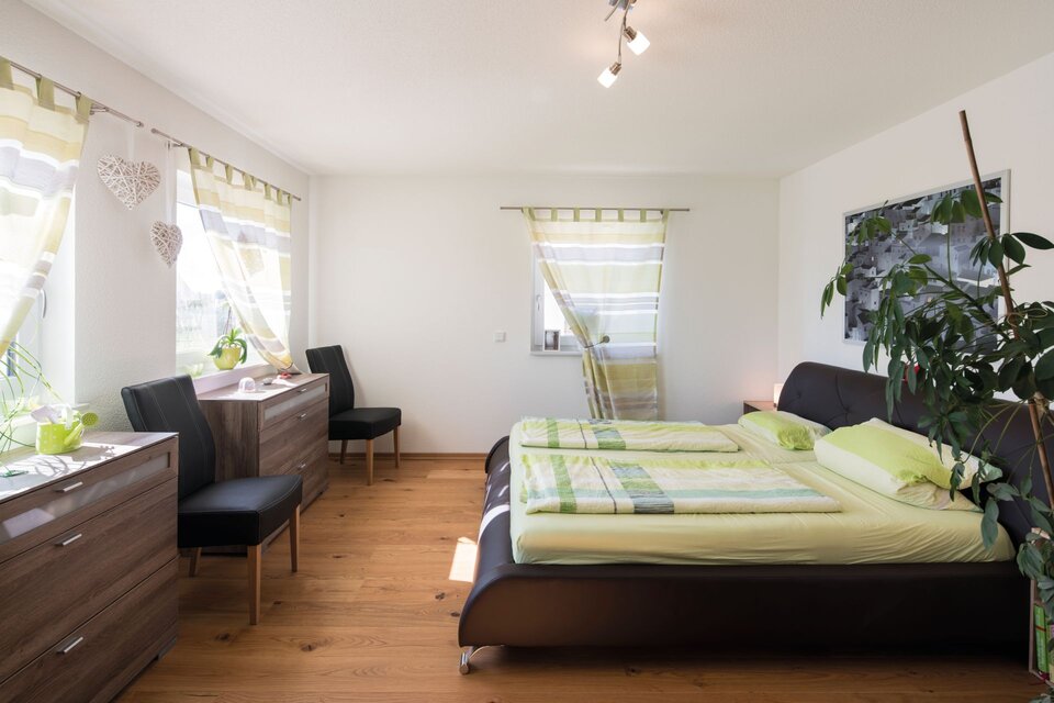 Helles Schlafzimmer im FingerHaus MEDLEY 310 mit großem Doppelbett, Holzfußboden, Sideboards und grün-weiß gestreiften Vorhängen.
