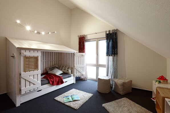 Kinderzimmer mit weißem Holzbett im Hüttenstil, dunklem Teppichboden und schräger Decke. Vor dem Bett liegen zwei Teppiche, ein offenes Buch und Sitzhocker. Durch das große Fenster fällt Tageslicht in den Raum.