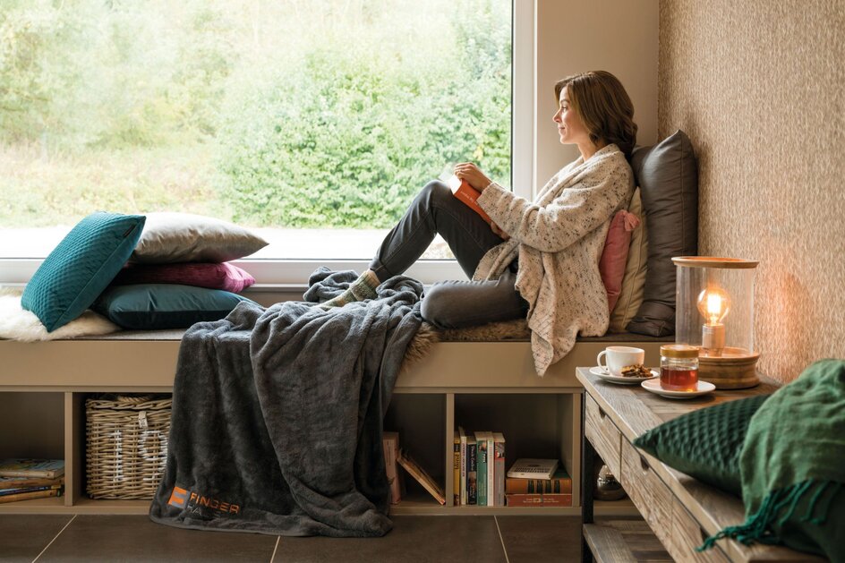 Eine Frau sitzt gemütlich auf einer Fensterbank mit Decke, Kissen und einem Buch, während neben ihr Tee und Gebäck stehen – ein behaglicher Moment im FingerHaus SENTO.