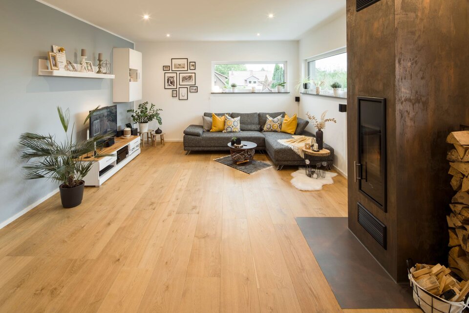 Wohnzimmer mit grauer Couch, Fernseherwand und Kamin mit Holzlager. Holzfußboden, Pflanzen und warme Beleuchtung schaffen eine gemütliche Atmosphäre.