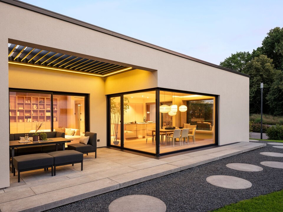 FingerHaus Bungalow NIVO in Frankenberg mit großer Eckverglasung, beleuchtetem Essbereich und überdachter Terrasse mit Sitzmöbeln.