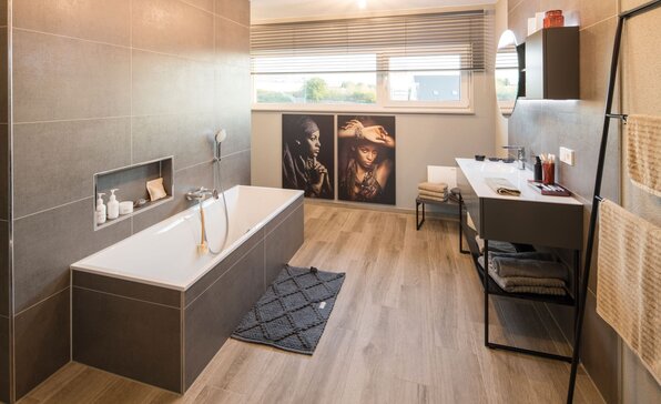 Modernes Badezimmer im Musterhaus MEDLEY 300 B in Kassel mit rechteckiger Badewanne, grauen Wandfliesen, Holzoptik-Boden und stilvoller Dekoration.