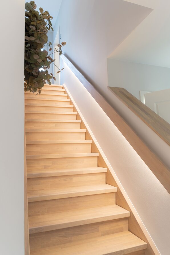 Helle geschlossene Holztreppe mit integrierter Wandbeleuchtung und schlichtem Handlauf. Weiß gestrichene Wände und modernes, minimalistisches Design betonen die klare Linienführung im Musterhaus SENTO Bad Vilbel.