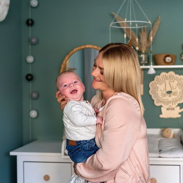 Eine Frau hält ein Baby im Arm in einem liebevoll eingerichteten Kinderzimmer mit grüner Wand, Wanddeko und Wickelkommode.