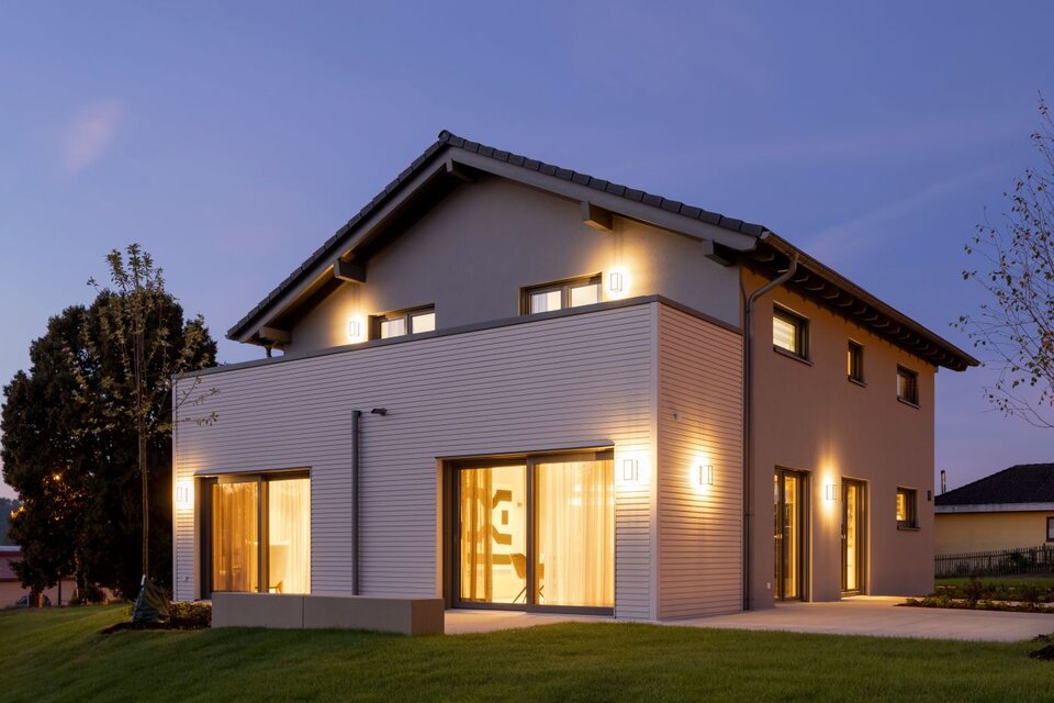 Fertighaus FingerHaus ARTIS 300 in Würzburg bei Abenddämmerung mit beleuchteter Fassade, Satteldach und großflächigen Fenstern, umgeben von gepflegtem Rasen.